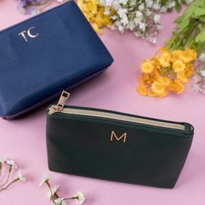 May include: Two zippered pouches, one navy blue with the initials "TC" and one dark green with the initial "M" in gold lettering. The pouches are on a pink background with white flowers.