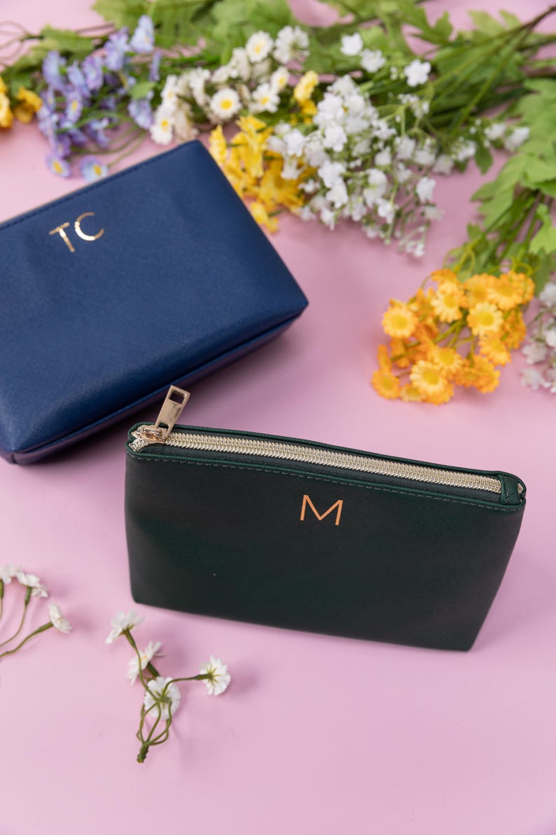 May include: Two zippered pouches, one navy blue with the initials "TC" and one dark green with the initial "M" in gold lettering. The pouches are on a pink background with white flowers.