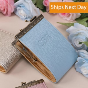May include: A light blue wallet with a textured surface and the embossed letters CSR. It features a bronze-toned metal kiss-lock clasp with intricate detailing and an open side revealing a gold-colored interior lining with visible card slots.