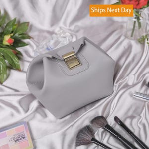 May include: A light gray pouch with a structured silhouette and a gold-toned metal clasp closure on the front. The clasp features a small rectangular plate with the engraved text Megan. The pouch has a smooth, matte finish and stitched detailing.