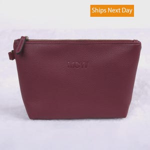 May include: A burgundy zippered pouch with a pebbled texture and the letters MCW embossed in the center. The pouch features a gold-toned metal zipper closure along the top edge and a small matching side tab attached to the zipper pull hardware.