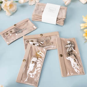 May include: Beige jewelry pouches with clear windows, showcasing necklaces and a brooch. The pouches are arranged on a light blue surface, with a stack of pouches and floral accents. The pouches have snap closures.