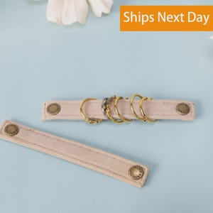 May include: Two beige suede-like fabric strips with metallic snap buttons at each end. One strip holds five small circular metal rings in gold and silver tones. The buttons are embossed with the text "ANGELINAS PALACE".