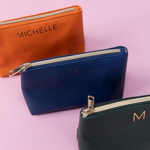 May include: Three zippered pouches in different colors. The top pouch is orange with the name "Michelle" printed on it. The middle pouch is blue with the initials "TC" printed on it. The bottom pouch is green with the initial "M" printed on it.