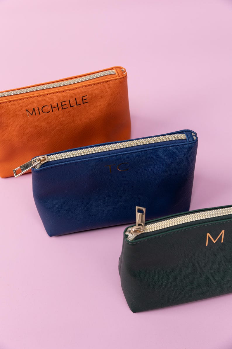 May include: Three zippered pouches in different colors. The top pouch is orange with the name "Michelle" printed on it. The middle pouch is blue with the initials "TC" printed on it. The bottom pouch is green with the initial "M" printed on it.