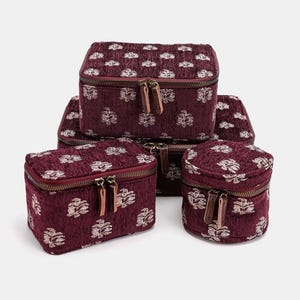 May include: A set of burgundy fabric cosmetic bags in various sizes. Each bag features a repeating floral pattern in a lighter color, a zipper closure, and leather zipper pulls. The bags are arranged in a group.