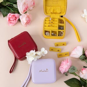 May include: Three small square jewelry boxes with a zipper closure and a wrist strap. The boxes are yellow, red, and purple. The boxes are labeled "The Gift", "Caroline", and "Madison".