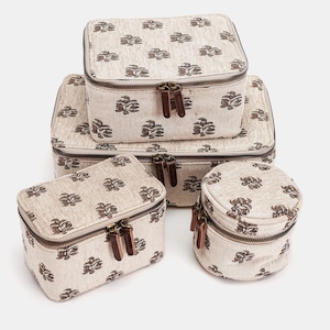 May include: A set of five beige fabric storage boxes with a repeating brown floral pattern. Each box has a zipper closure with brown leather pulls. The boxes are various sizes, including rectangular and round shapes.