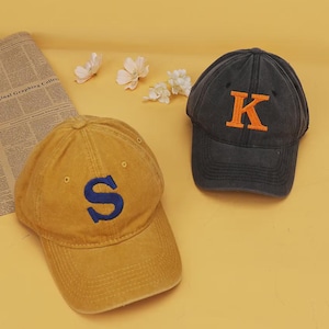May include: Two baseball caps, one mustard yellow with a navy blue embroidered letter S and one charcoal gray with an orange embroidered letter K. Both caps feature a six-panel construction with a curved brim and small ventilation eyelets.