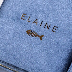 K&ouml;nnte beinhalten: Eine kleine, blaue Schmuckschatulle aus Samt mit einem goldenen Rei&szlig;verschluss. Die Schachtel tr&auml;gt den Namen "ELAINE" und ein goldenes Fischskelett-Design auf der Vorderseite.