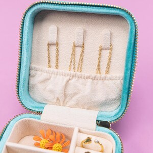 K&ouml;nnte beinhalten: Eine Schmuckschatulle aus t&uuml;rkisfarbenem Samt mit Rei&szlig;verschluss. Die Box hat F&auml;cher f&uuml;r Ringe, Ohrringe und Halsketten. Die Box ist ge&ouml;ffnet und das Innere ist mit wei&szlig;em Samt ausgekleidet.