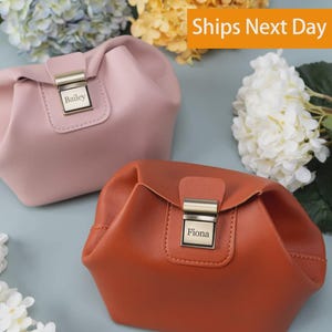 May include: Two structured pouches, one in light pink and one in burnt orange, each featuring a rectangular metal clasp closure. The pink pouch is engraved with the name Bailey, and the orange pouch is engraved with the name Fiona on the metal hardware.