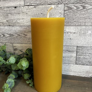 May include: A golden yellow pillar candle with a white wick. The candle is cylindrical and stands upright. The background is a light-colored wooden surface with green foliage.