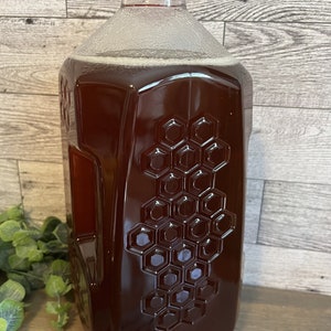 May include: A large, clear plastic bottle filled with honey. The bottle has a red lid and a honeycomb pattern embossed on the side.