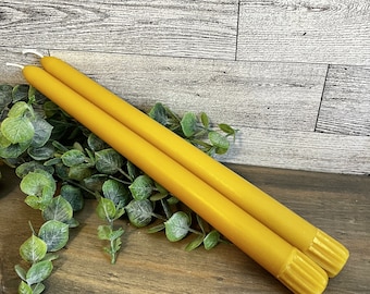 May include: Two yellow beeswax candles with a textured, ridged top. The candles are on a wooden surface with green foliage.