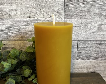 May include: A large, golden yellow pillar candle with a white wick. The candle is sitting on a wooden surface.