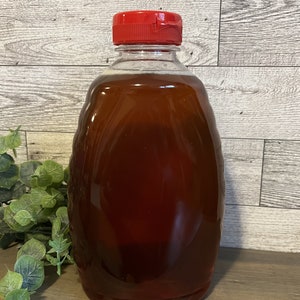 May include: A clear glass bottle with a red lid filled with honey. The bottle is sitting on a wooden surface.