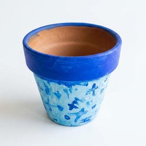 May include: A terracotta plant pot featuring a solid vibrant blue painted rim and a light blue base decorated with small, dark blue hand-painted cross-shaped marks. The interior of the pot remains unpainted, showing the natural brown clay color.