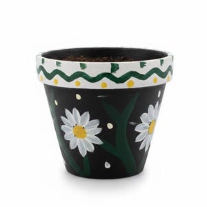 May include: A black ceramic flower pot with a white rim featuring a green and yellow wavy design. The pot is decorated with white daisies with yellow centers and green stems. The pot is filled with soil.