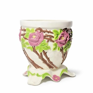 May include: A white ceramic planter featuring a raised relief design of pink roses, green leaves, and brown thorny stems. The vessel stands on a flared base with four feet, each decorated with a small pink flower and green accents.