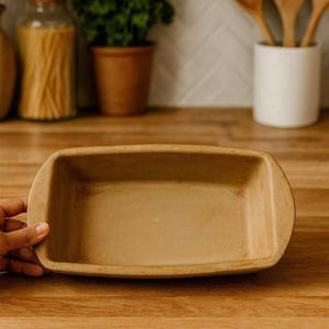 May include: A rectangular, tan-colored ceramic loaf pan with rounded edges. The baking dish is empty and appears to be new. The pan is sitting on a wooden surface. Kitchenware.
