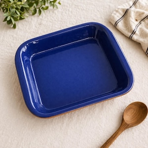 May include: A rectangular, cobalt blue ceramic baking dish with rounded corners. The dish has a glossy finish and a light brown base. A wooden spoon rests on the surface next to the dish. A beige and black striped cloth is in the background.