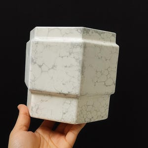 May include: A white ceramic planter with a faceted, geometric shape and a grey marbled pattern. The vessel features a stepped design with a wider top section and a narrower base, showcasing a distinct network of grey veins across the surface.