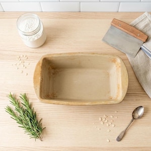 May include: A tan rectangular ceramic loaf pan sits on a light wood surface, surrounded by baking ingredients. A glass jar of flour, a metal scraper with a wooden handle, a sprig of rosemary, and a spoon are also present, suggesting a baking theme.
