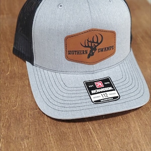 Op de afbeelding: Een grijze en zwarte truckerpet met een bruine, leerachtige patch aan de voorzijde met een zwarte afbeelding van een hertengewei en de tekst SOUTHERN SWAMPS. Een zwart-witte sticker op de klep vermeldt RICHARDSON 112 TRUCKER ORIGINAL ADJUSTABLE.