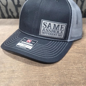 Può includere: Cappellino da camionista nero e grigio con la scritta "SAME ASSHOLE DIFFERENT DAY" su una toppa quadrata. Il cappellino ha una visiera curva e un retro in rete. L'etichetta del marchio "RICHARDSON" è visibile.