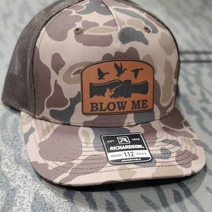 Pode incluir: Um chapéu de caminhoneiro marrom e camuflado com um remendo de couro marrom que diz "Blow Me" com um design de pato e chamada de pato. O chapéu é ajustável e tem o logotipo da Richardson na parte de trás.