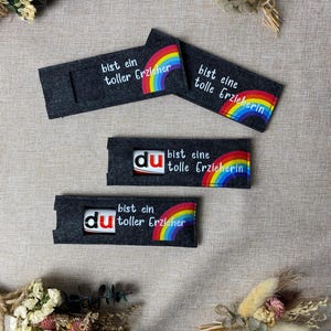 Può includere: Quattro portaoggetti in feltro grigio scuro con dettagli arcobaleno e testo. Il testo include frasi come "bist ein toller Erzieher" e "bist eine tolle Erzieherin". I portaoggetti sono disposti su un tessuto dai toni neutri, con composizioni di fiori secchi.