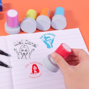 Pode incluir: Uma coleção de carimbos coloridos com bases cinzentas. Um carimbo está a ser usado para imprimir "Mrs. Sophia says Great work!" em papel pautado. Outros carimbos incluem "Well Done!" e "You are Amazing!" em várias cores.