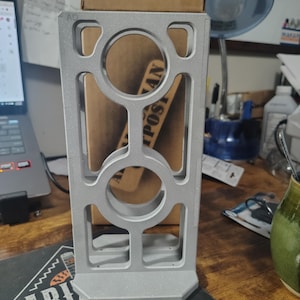 May include: A silver-colored, 3D-printed vertical stand featuring a rectangular frame with two circular cutouts stacked vertically. The structure has a flat base and a textured, matte finish, with the letters AQ embossed in the top left corner.