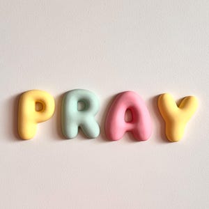 May include: Four individual, rounded, three-dimensional letters spelling the word PRAY. The letter P is yellow, the letter R is light mint green, the letter A is soft pink, and the letter Y is yellow. Each letter has a smooth, matte finish.