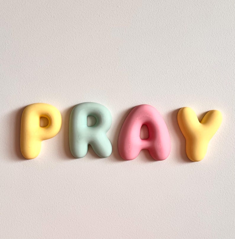 May include: Four individual, rounded, three-dimensional letters spelling the word PRAY. The letter P is yellow, the letter R is light mint green, the letter A is soft pink, and the letter Y is yellow. Each letter has a smooth, matte finish.