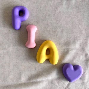 May include: A set of four small, smooth, matte-finish objects arranged diagonally. The collection includes a purple letter P, a light pink bone-shaped piece, a yellow letter A, and a purple heart-shaped object, all featuring rounded edges.