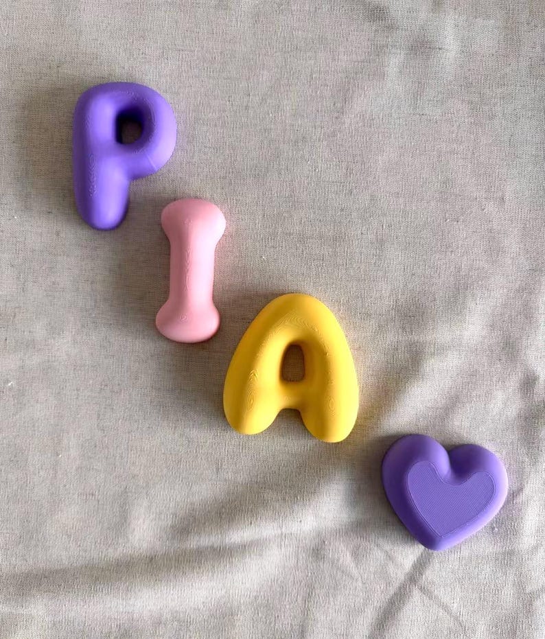 May include: A set of four small, smooth, matte-finish objects arranged diagonally. The collection includes a purple letter P, a light pink bone-shaped piece, a yellow letter A, and a purple heart-shaped object, all featuring rounded edges.