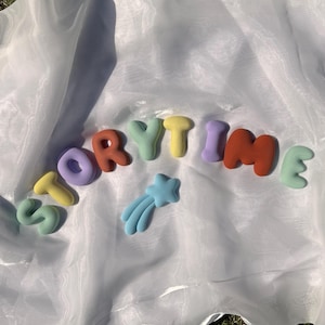 Puede incluir: Un conjunto de letras individuales, redondeadas y de colores pastel que forman la palabra STORYTIME, acompañadas de una pequeña forma de estrella fugaz de color azul claro. Las letras son de color verde menta, amarillo, lavanda y naranja terracota.