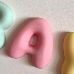May include: A three-dimensional letter A in a soft matte pink finish with a rounded, inflated appearance. The letter is positioned between a partial light mint green shape on the left and a partial yellow shape on the right against a white surface.