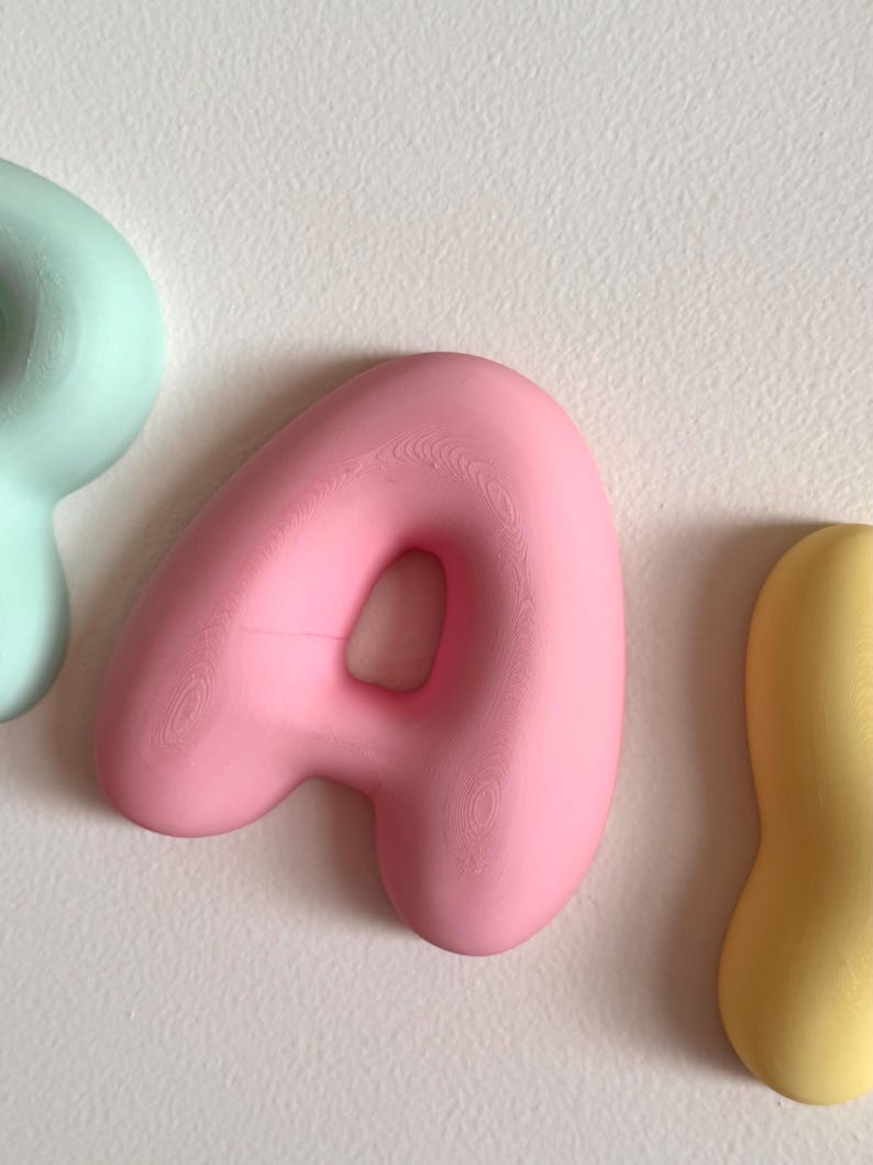 May include: A three-dimensional letter A in a soft matte pink finish with a rounded, inflated appearance. The letter is positioned between a partial light mint green shape on the left and a partial yellow shape on the right against a white surface.