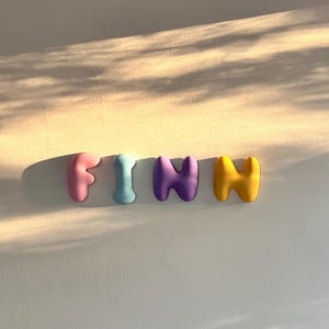 May include: Four individual three-dimensional letters spelling the name FINN. The letter F is light pink, the letter I is light blue, the first N is purple, and the second N is yellow. Each letter has a smooth, rounded, bubble-like shape.