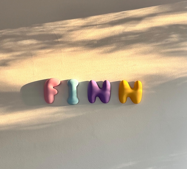 May include: Four individual three-dimensional letters spelling the name FINN. The letter F is light pink, the letter I is light blue, the first N is purple, and the second N is yellow. Each letter has a smooth, rounded, bubble-like shape.