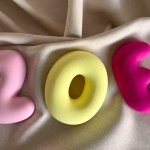 May include: Three-dimensional letters spelling the name ZOE. The letter Z is light pink, the letter O is a bright yellow ring shape, and the letter E is a vibrant magenta. Each letter has a smooth, rounded, and puffy appearance with a matte finish.