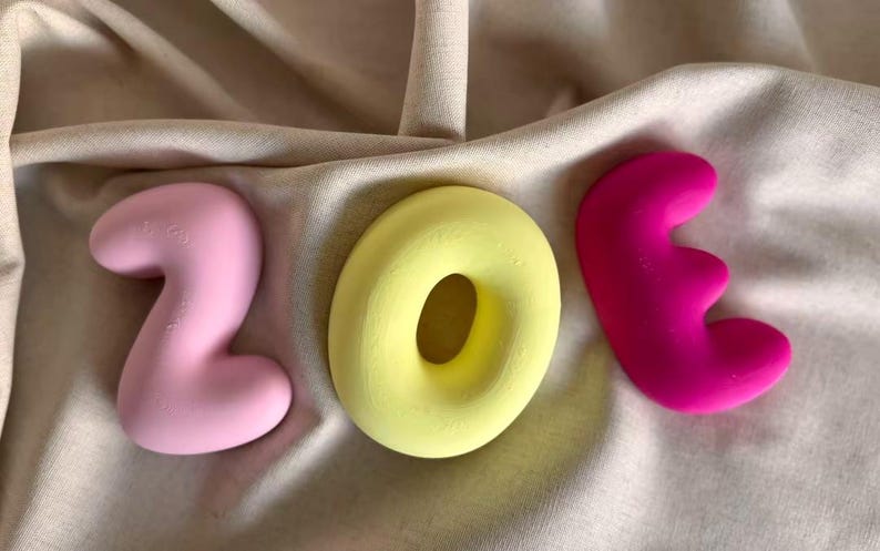 May include: Three-dimensional letters spelling the name ZOE. The letter Z is light pink, the letter O is a bright yellow ring shape, and the letter E is a vibrant magenta. Each letter has a smooth, rounded, and puffy appearance with a matte finish.