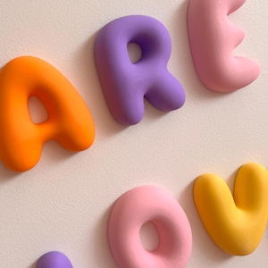 May include: A set of three-dimensional wall-mounted letters spelling out the word AREOV. The letters feature a smooth, rounded, puffy design in vibrant colors including orange, purple, light pink, and yellow, arranged against a plain wall.