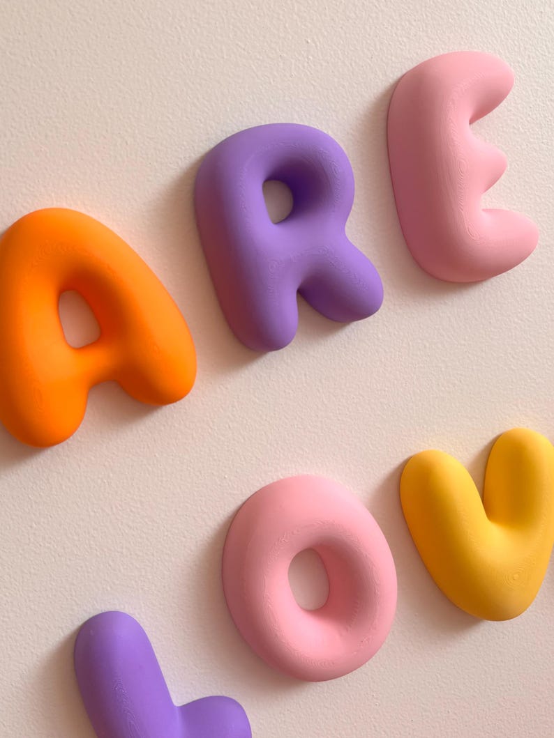 May include: A set of three-dimensional wall-mounted letters spelling out the word AREOV. The letters feature a smooth, rounded, puffy design in vibrant colors including orange, purple, light pink, and yellow, arranged against a plain wall.