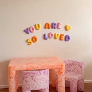 May include: A rectangular table and two chairs with a speckled orange and purple pattern, accompanied by wall-mounted, three-dimensional, multi-colored letters and a heart shape that spell out the phrase YOU ARE SO LOVED in a rounded font.