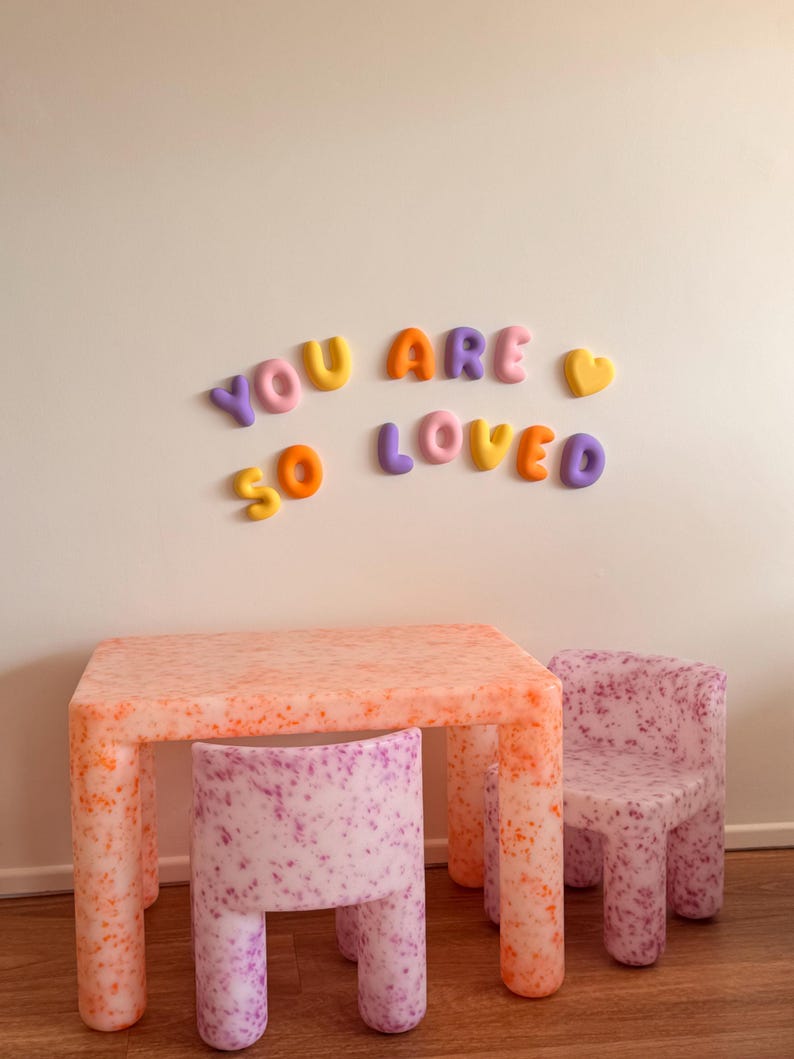 May include: A rectangular table and two chairs with a speckled orange and purple pattern, accompanied by wall-mounted, three-dimensional, multi-colored letters and a heart shape that spell out the phrase YOU ARE SO LOVED in a rounded font.