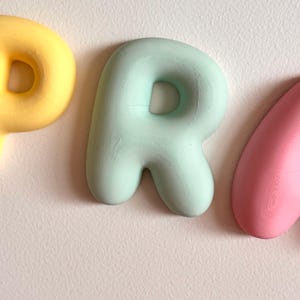 May include: A three-dimensional letter P in a soft mint green color with a rounded, bubble-like shape. The letter is positioned between a partial yellow letter to the left and a partial pink letter to the right, all featuring a smooth finish.